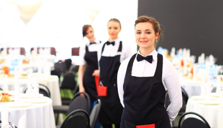 Three professional catering staff members in black aprons and bow ties standing in a banquet hall with round tables set for an event, with one server in focus and two blurred in the background.