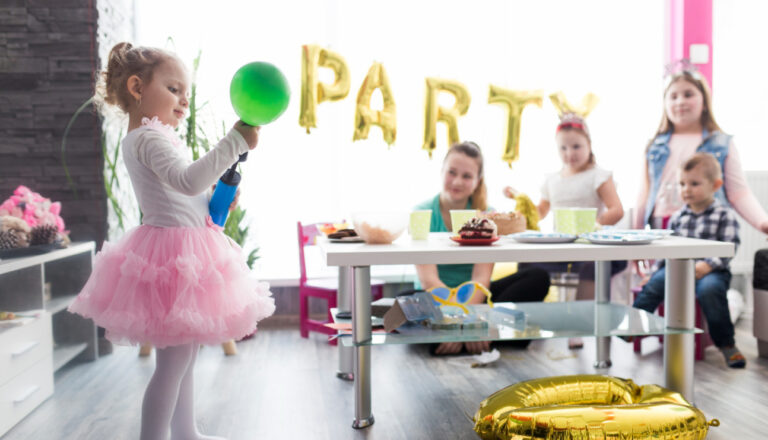 Young girl in a pink tutu holding a green balloon at a children’s birthday party, with other kids and an adult gathered around a table with cake and snacks, and gold “PARTY” balloons in the background.