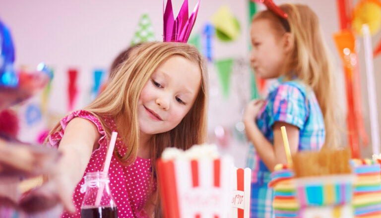 Smiling young girl wearing a pink paper crown reaches across a colorful birthday party table with drinks, striped popcorn boxes, and decorations, while another child stands in the background.