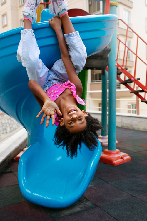 Smiling child hanging upside down on a playground slide, enjoying outdoor playtime